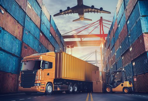 Verschiedene Transportfahrzeuge stehen im Hafen zwischen Seecontainern, über ihnen fliegt ein Flugzeug.