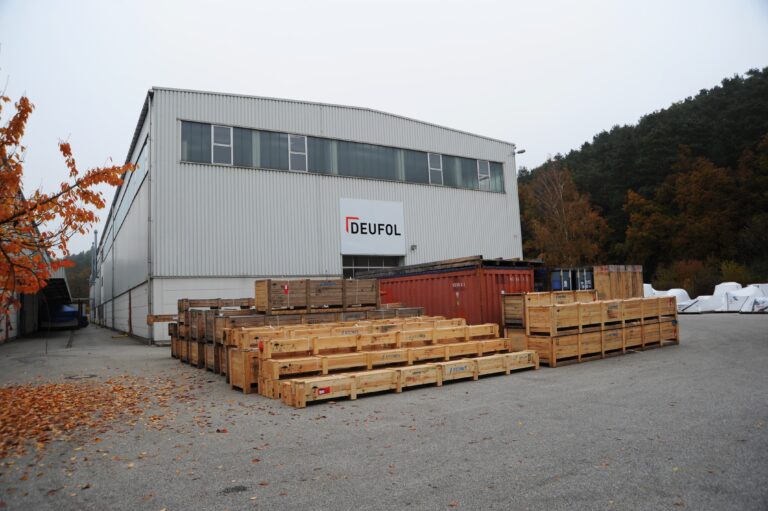 Transportpaletten neben Containern vor einer DEUFOL-Lagerhalle gestapelt.