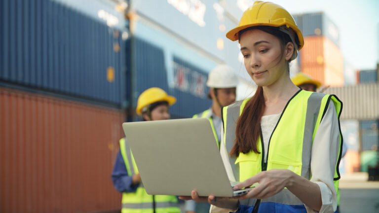 Eine Arbeiterin mit Helm und Warnweste steht vor Frachtcontainern und überprüft mit dem CPC-Tracker auf ihrem Laptop den Transport der Industriegüter.